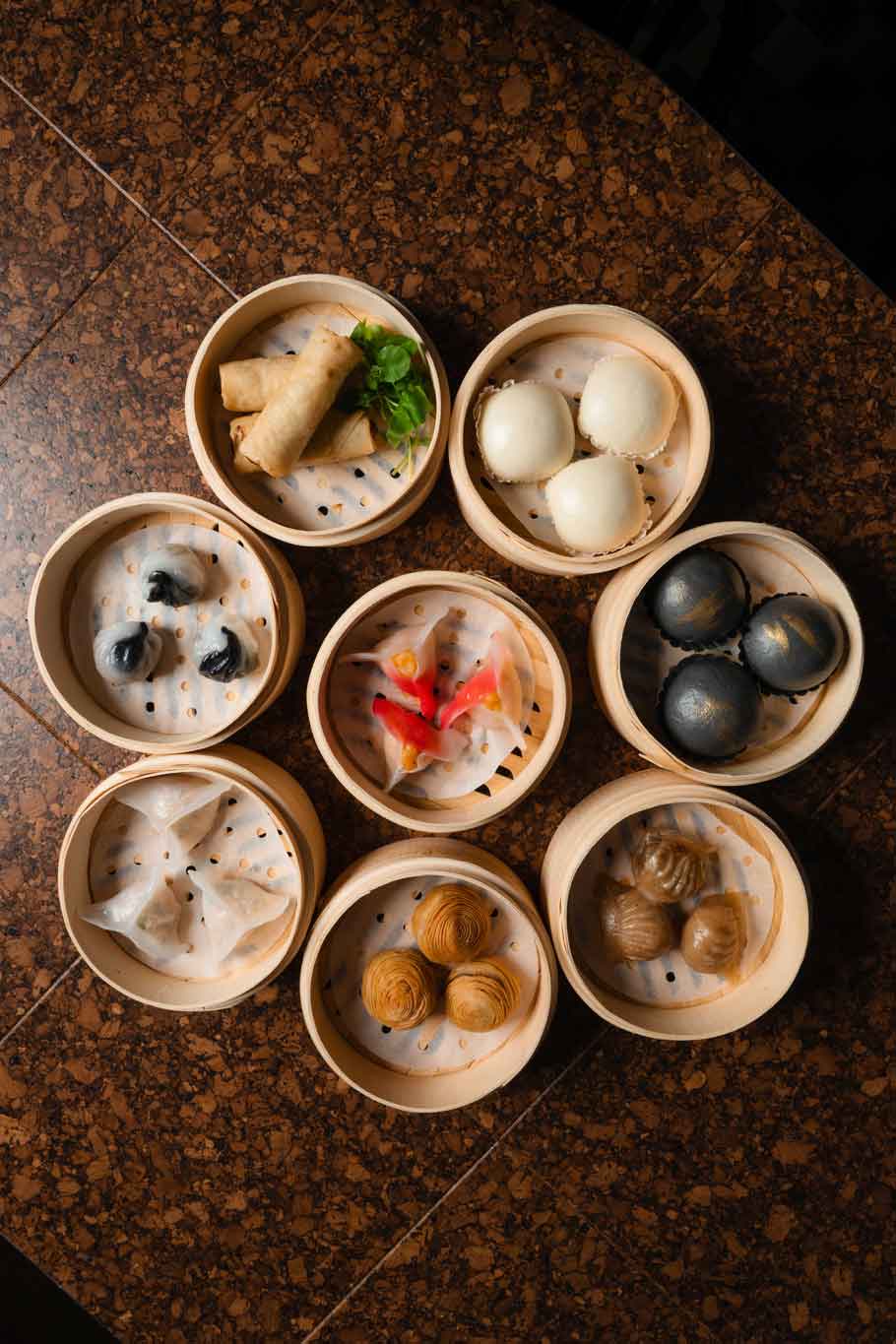Assorted dumplings arranged in round bamboo steamers on a cork tabletop.