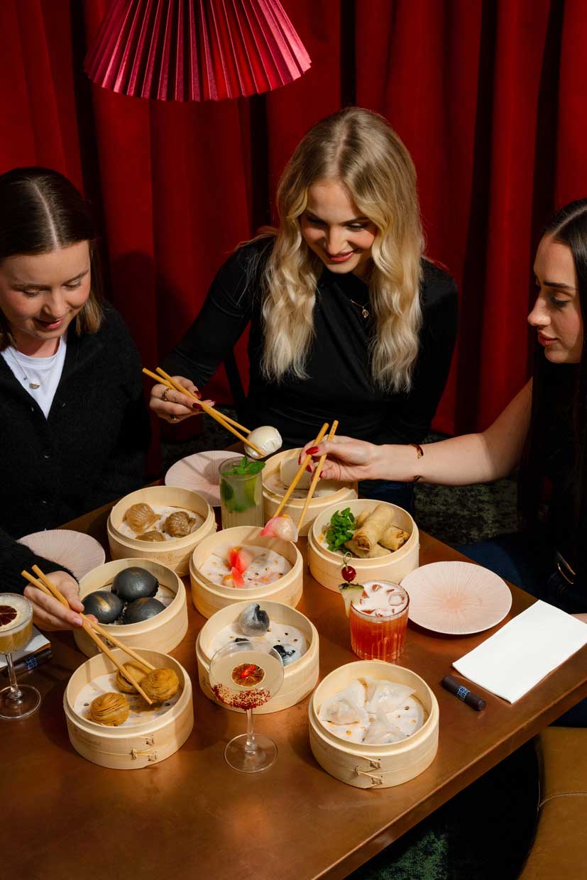 Three women smiling and sharing a colourful spread of dumplings, cocktails, and small plates.