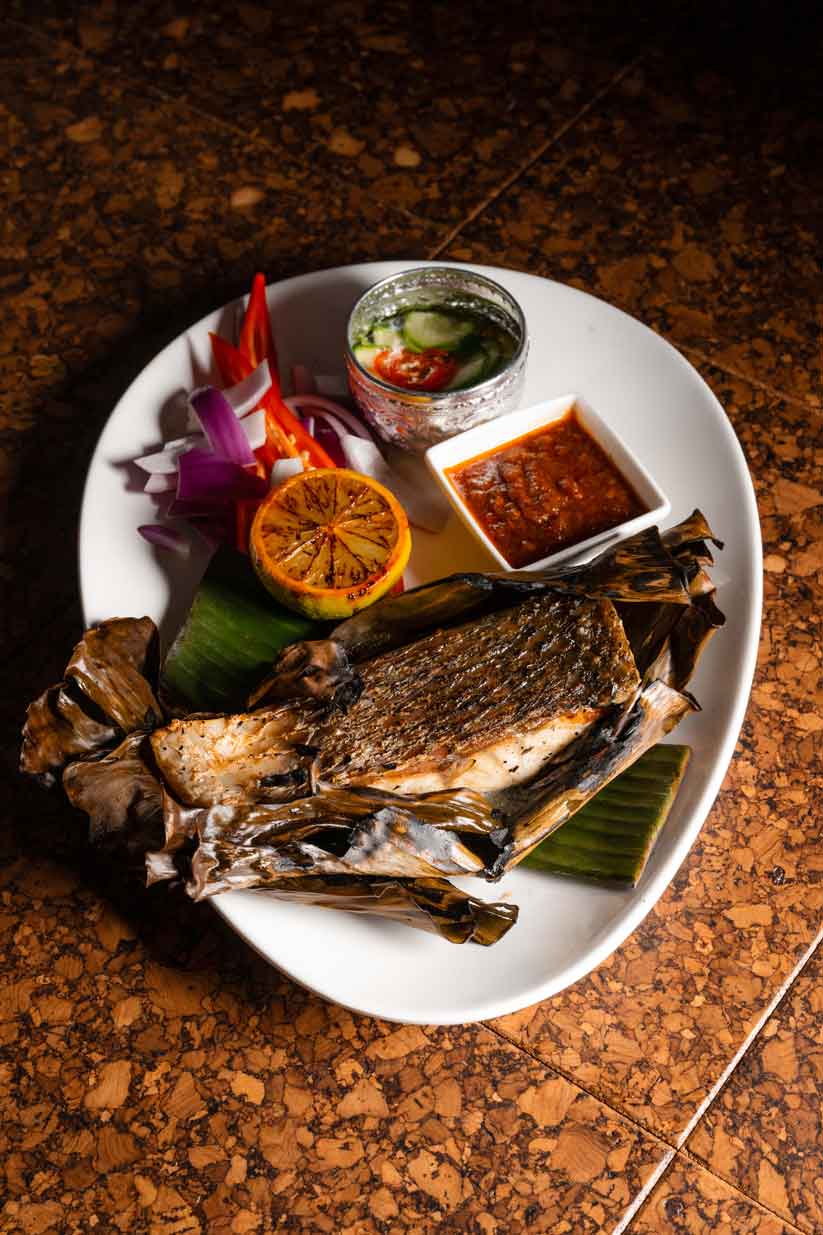 Whole grilled fish served with lime, herbs, red onion, dipping sauce, and sambal on a ceramic plate.