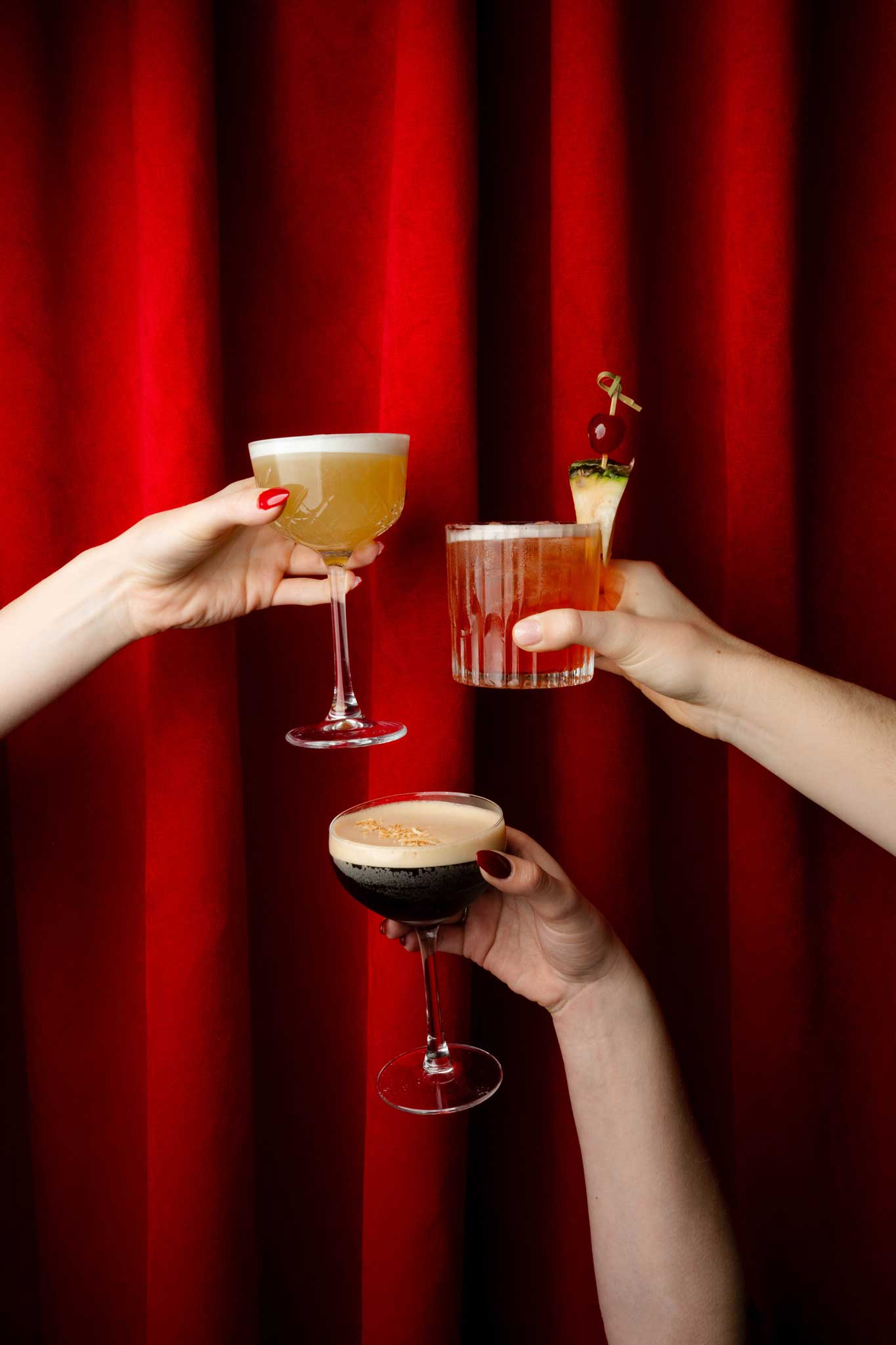 Three hands raising cocktails in front of a dramatic red velvet curtain — a yellow martini, a dark espresso martini, and a vibrant tropical drink with pineapple garnish.