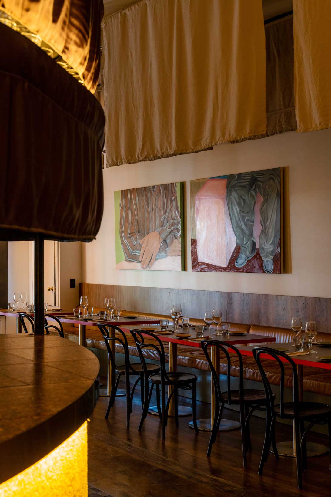 Warmly lit dining area with wooden tables, black chairs, hanging fabric panels, and contemporary paintings on the wall, creating an intimate and artistic atmosphere.